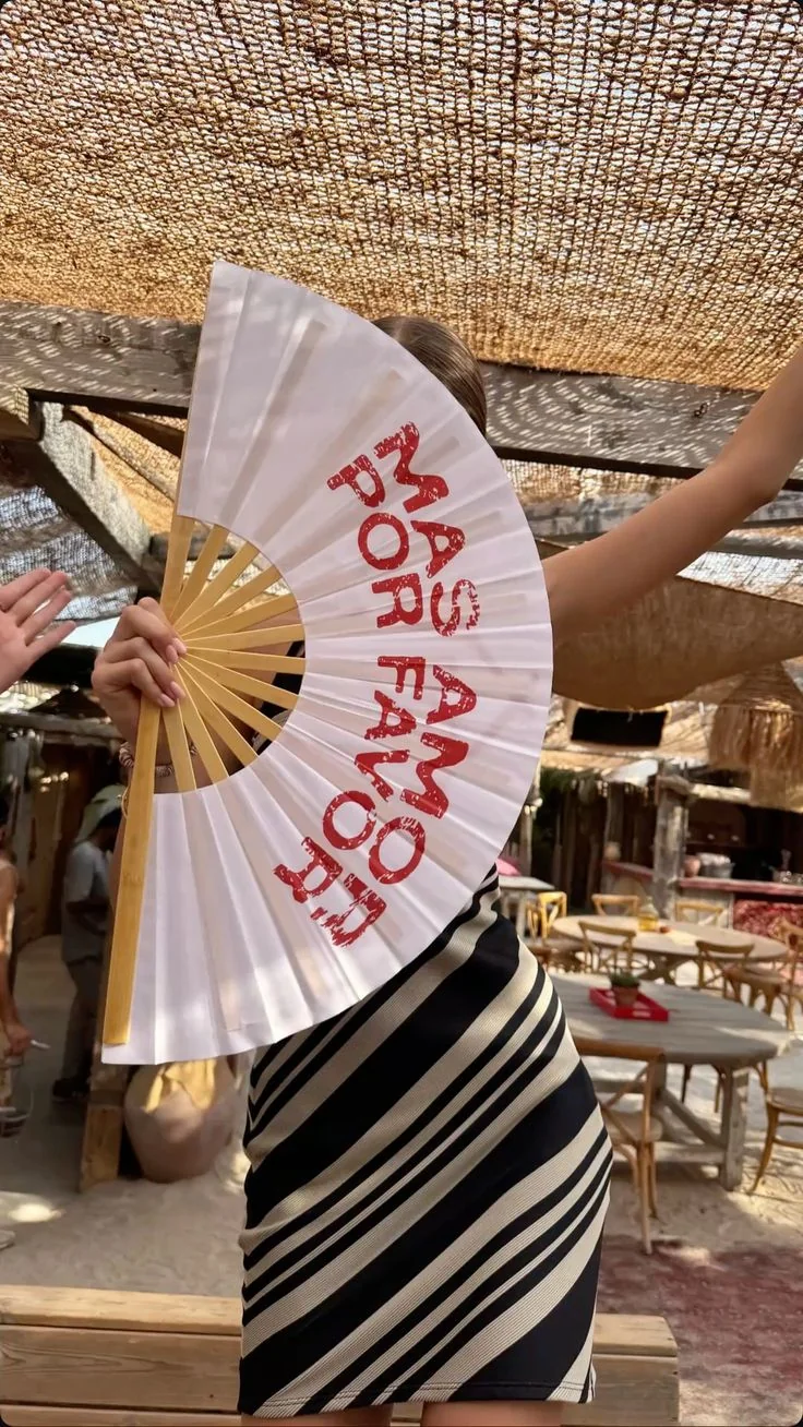 Abanico blanco personalizado con texto rojo, sostenido frente a una terraza